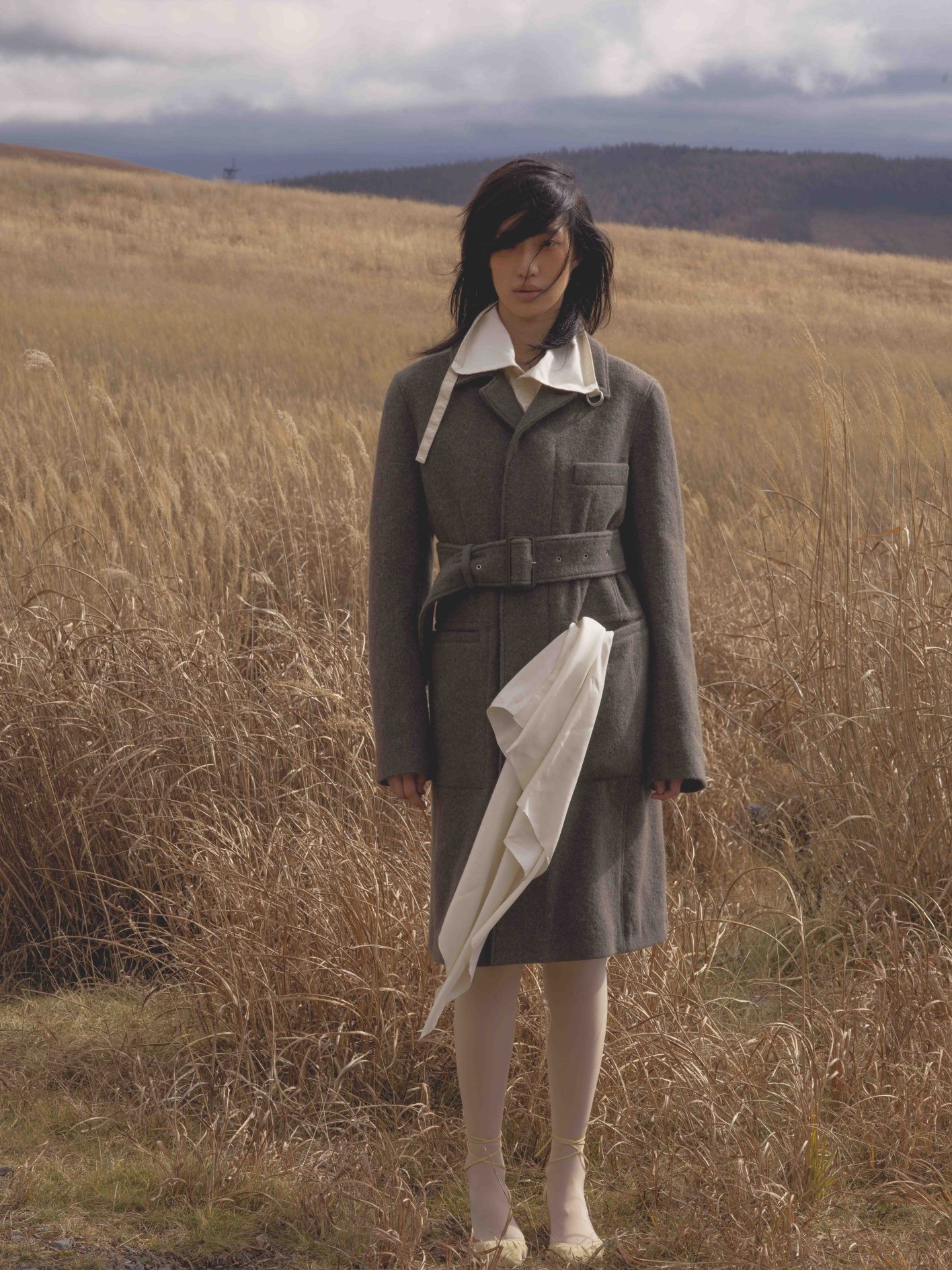「自然から抽出したような中間色がいつも魅力的な〈ルメール〉。深く落ち着いたカーキ色に、静かな冬の森を想いました」（井伊さん）。「『枯野見』と称して、わざわざ枯れた野原を鑑賞する江戸っ子の余裕とセンスに触発されて、頰や唇にも侘びた色の遊びをほんのりと」（UDAさん）。マニッシュなベルト付きコート。首元からシャツの大きな襟を覗かせて。コート¥280,000、シャツ ¥122,000、シューズ ヒール5.5cm ¥109,000＊参考価格（以上ルメール | エドストローム オフィス）／スカーフ、タイツ＊スタイリスト私物