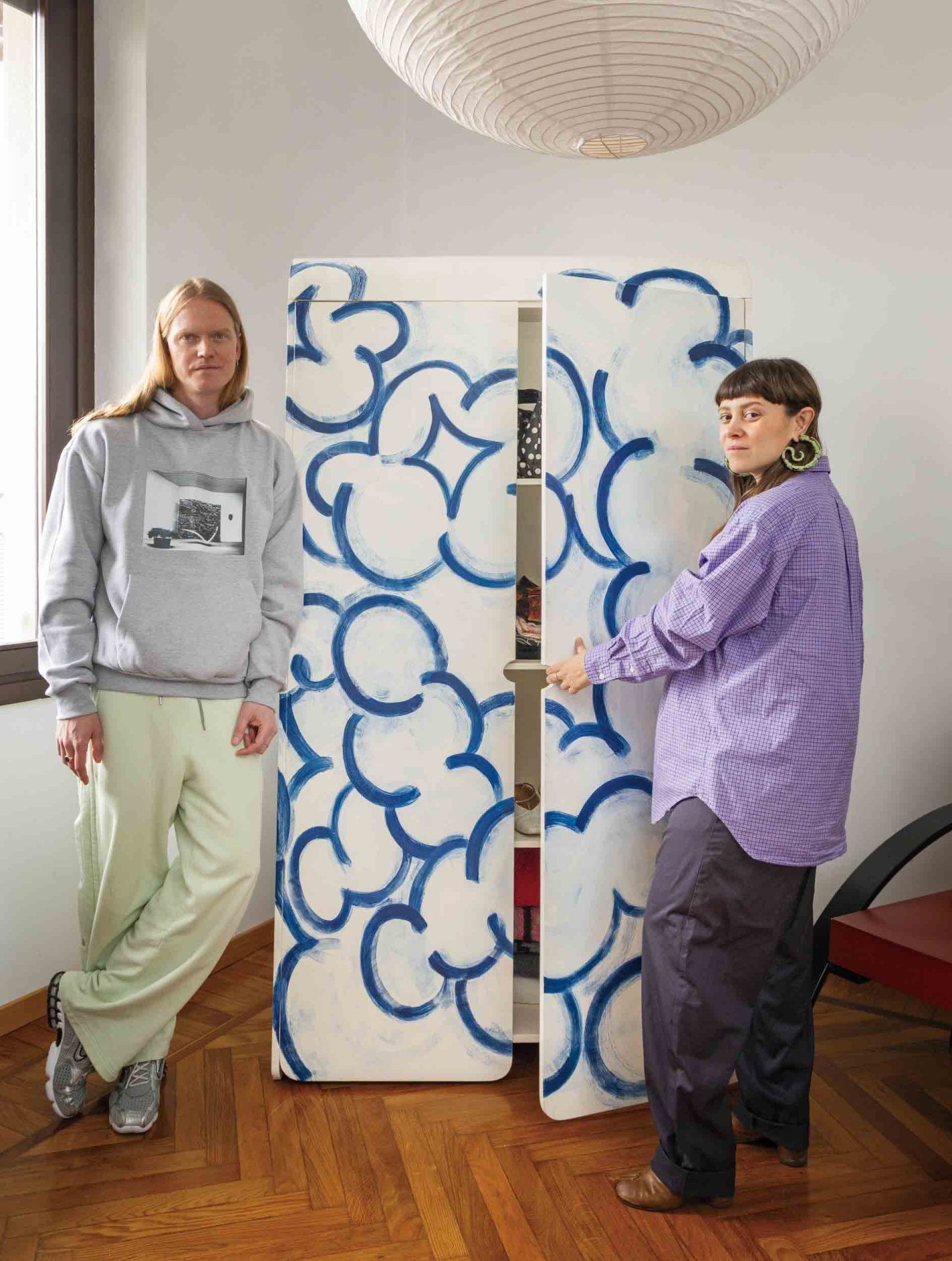 左から〈OLDER Studio〉ファウンダー Morten Thuesen、Letizia Caramia/自作のキャビネット〈ブクリコロ〉。雲のようなドローイングは、2023年のミラノ・デザインウィーク中に二人でライヴペインティングしたもので、収納を兼ねたアート作品でもある。レティツィアのシャツはヴィンテージの〈ラルフローレン〉、モーテンのパンツは〈オールダー〉と〈ファセッタズム〉のカプセルコレクション。